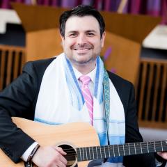 Man smiling and holding a guitar wearing a dark suit and tallit