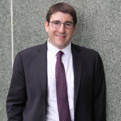 Man in grey suite wearing glasses against a wall