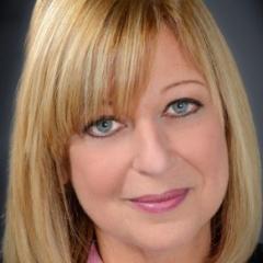 Cantor Jodi Schechtman: shoulder length blonde hair, blue eyes, smiling. Wearing pink lipstick and dark eyeliner. Wearing a pink blouse under a black blazer.