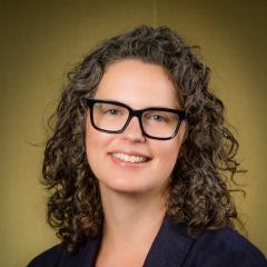 Brown curly hair, brown eyes, dark rimmed glasses. Smiling.