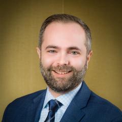 Face of cantor josh breitzer in a full trimmed beard. smiling and wearing a checked blue shirt