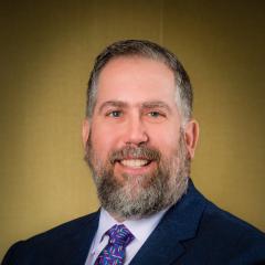 Smiling man with full grey-brown beard. Navy suite. Blue colorful tie.