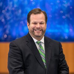 Cantor Seth Warner in a dark suit with a green tie and white shirt standing in front of a blue background smiling. Hands folded in front of him.