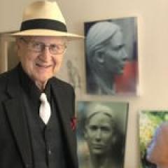Cantor Sheldon Merel, wearing a black shirt, white tie, and hat standing in front of photos and painting