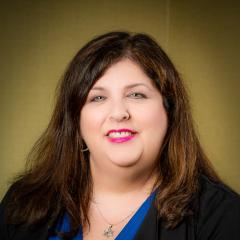 Smiling--wearing pink lipstick. Long brown hair. Blue shirt and black jacket.