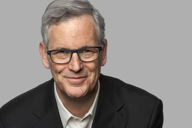 Man with grey hair and glasses smiling at the camera. Wearing a blue blazer and white collared shirt. 