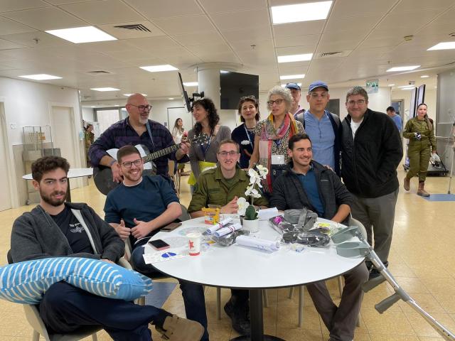 A group of IDF soldiers and several cantors standing and sitting around a table talking about the war