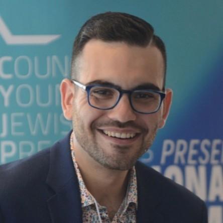 Smiling young man with short dark hair, wearing glasses, open collar multi-color shirt, and blue blazer