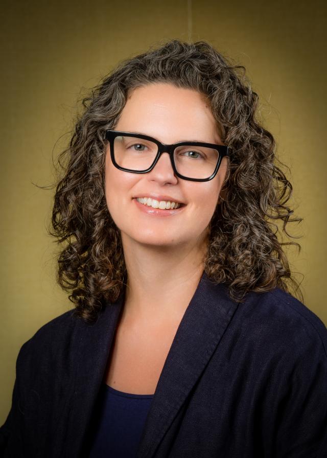 Brown curly hair, brown eyes, dark rimmed glasses. Smiling.